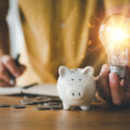 Mensch mit Glühbirne in der Hand einem Sparschwein auf dem Tisch und daneben geld
