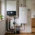Heizungsraum mit wandhängendem Gasbrennwertkessel und Innenmodul einer Luft-Wärmepumpe, Kupferrohre, digitales Thermostat, warmes Morgenlicht auf hellem Eichenboden.