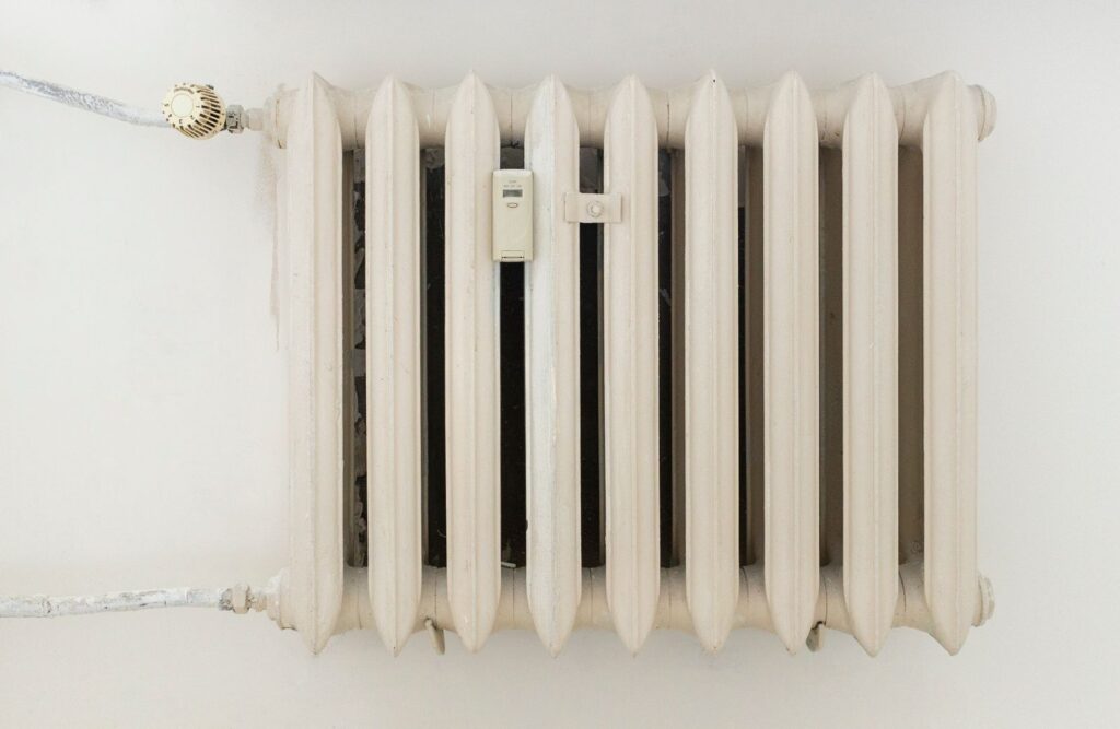 Weißer Gussheizkörper mit Heizkostenverteiler und Thermostatventil an einer hellen Wand, schmaler Spalt zur Wand